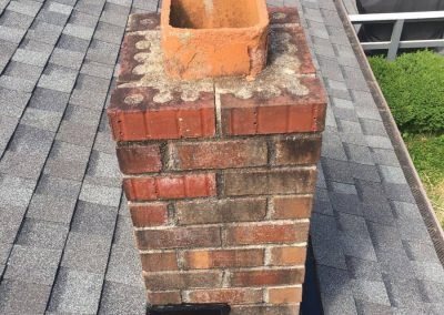 A red brick chimney on a gray shingled roof, viewed from above, with a patio visible in the background.