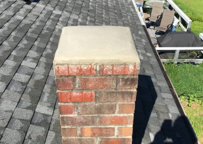 Brick chimney on a shingled roof with a concrete cap, surrounded by trees and patio furniture visible.