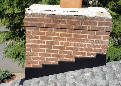 Brick chimney with a metal cap on a shingled roof, surrounded by green trees.
