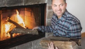 A person in a plaid shirt, like an expert from a Knowledge Center tutorial, places firewood into a lit stone fireplace, smiling warmly.