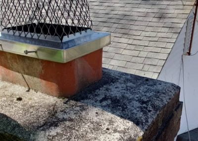 A metal chimney cap on a brick chimney, with shingled rooftops and trees in the background.