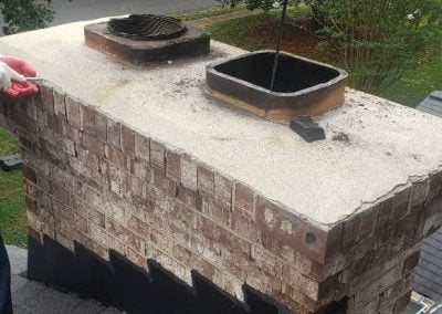 A brick chimney on a rooftop with tools and a coil rod visible, surrounded by trees.