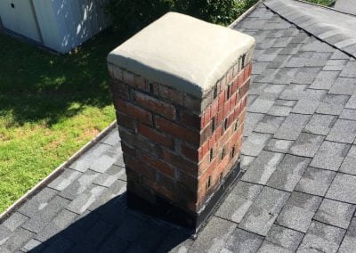 Brick chimney with a concrete cap on an asphalt shingle roof, with grass and sheds visible in the background.