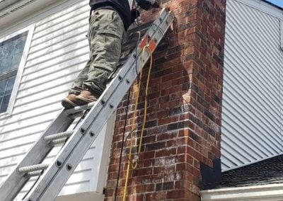 Person standing on a ladder drilling into a brick chimney attached to a white house.
