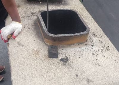 A person cleaning a chimney flue on a rooftop with tools and soot visible around the flue openings.