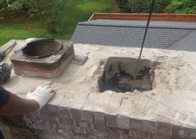 Person cleaning a brick chimney flue on a rooftop with a chimney brush and tools nearby.