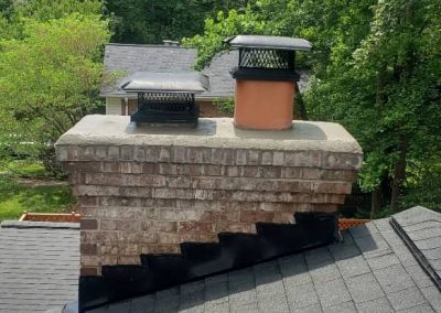 Brick chimney with two vents on a shingled roof, surrounded by trees and neighboring houses.