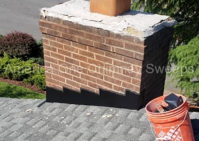 A brick chimney on a shingled roof with a Home Depot bucket nearby, surrounded by trees and street.