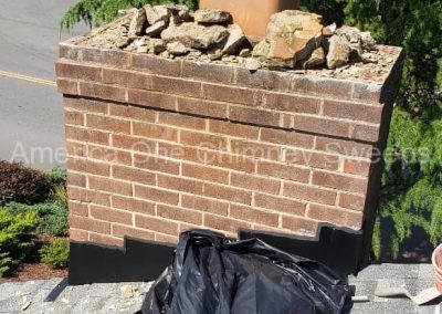 A brick chimney with rocks on top and a black plastic bag on a shingled roof, trees in the background.