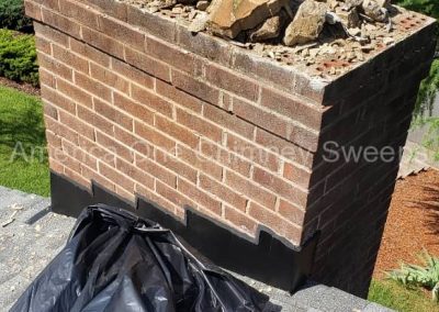 A brick chimney with loose rocks on top, a metal cap, a black trash bag, and an orange bucket nearby.