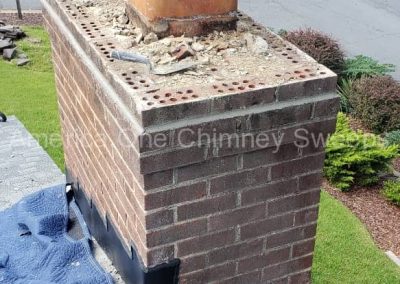 A brick chimney with loose debris on top, viewed from a rooftop next to a suburban street and yard.