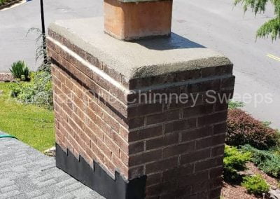 Brick chimney with a metal cap on a shingled roof, overlooking a suburban street with trees and houses.