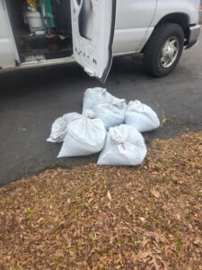 Six large white bags are on the ground beside an open van door on a paved surface near dried leaves.