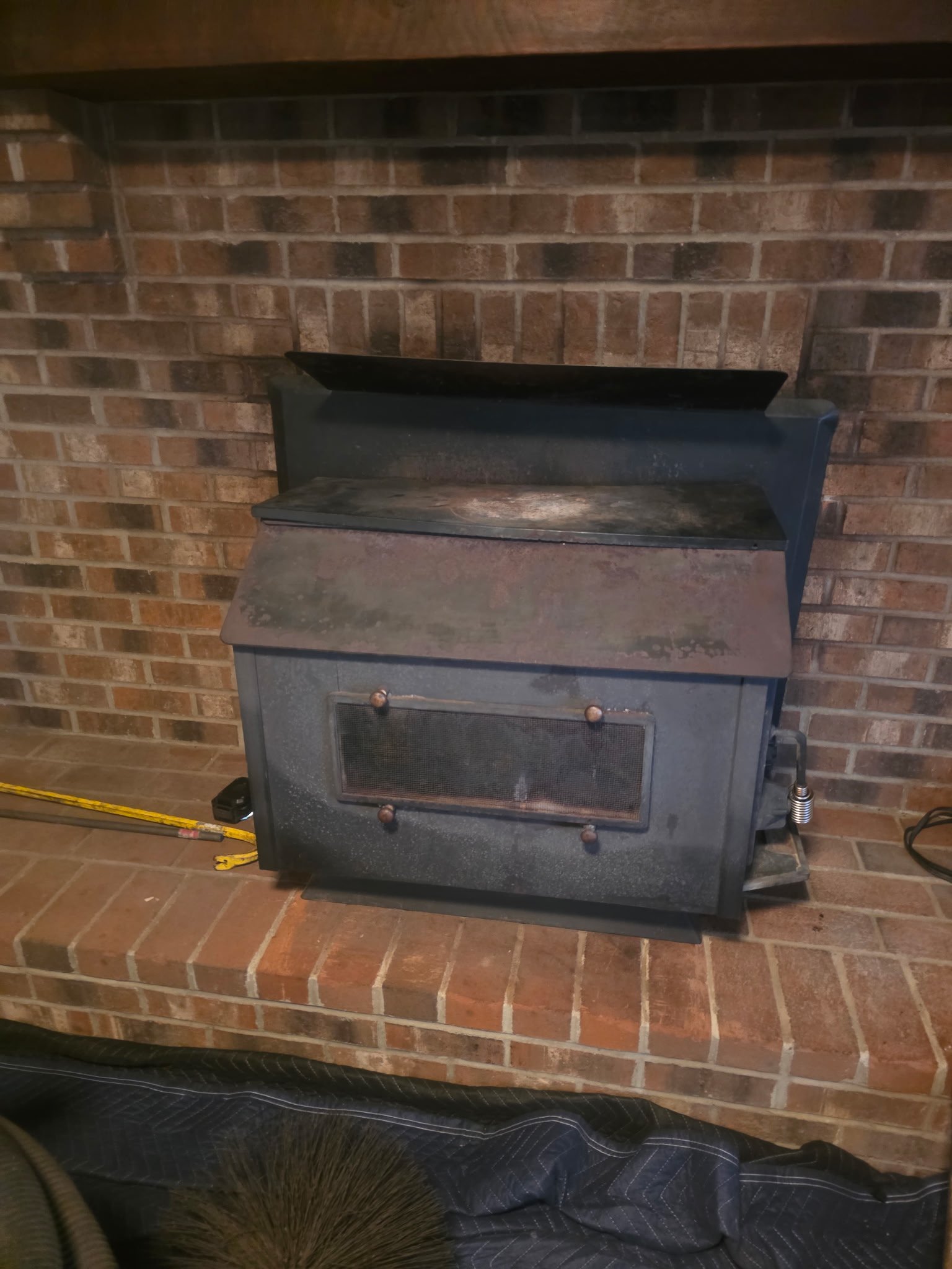 Black wood stove insert inside a brick fireplace, with a tape measure and cleaning brush nearby.